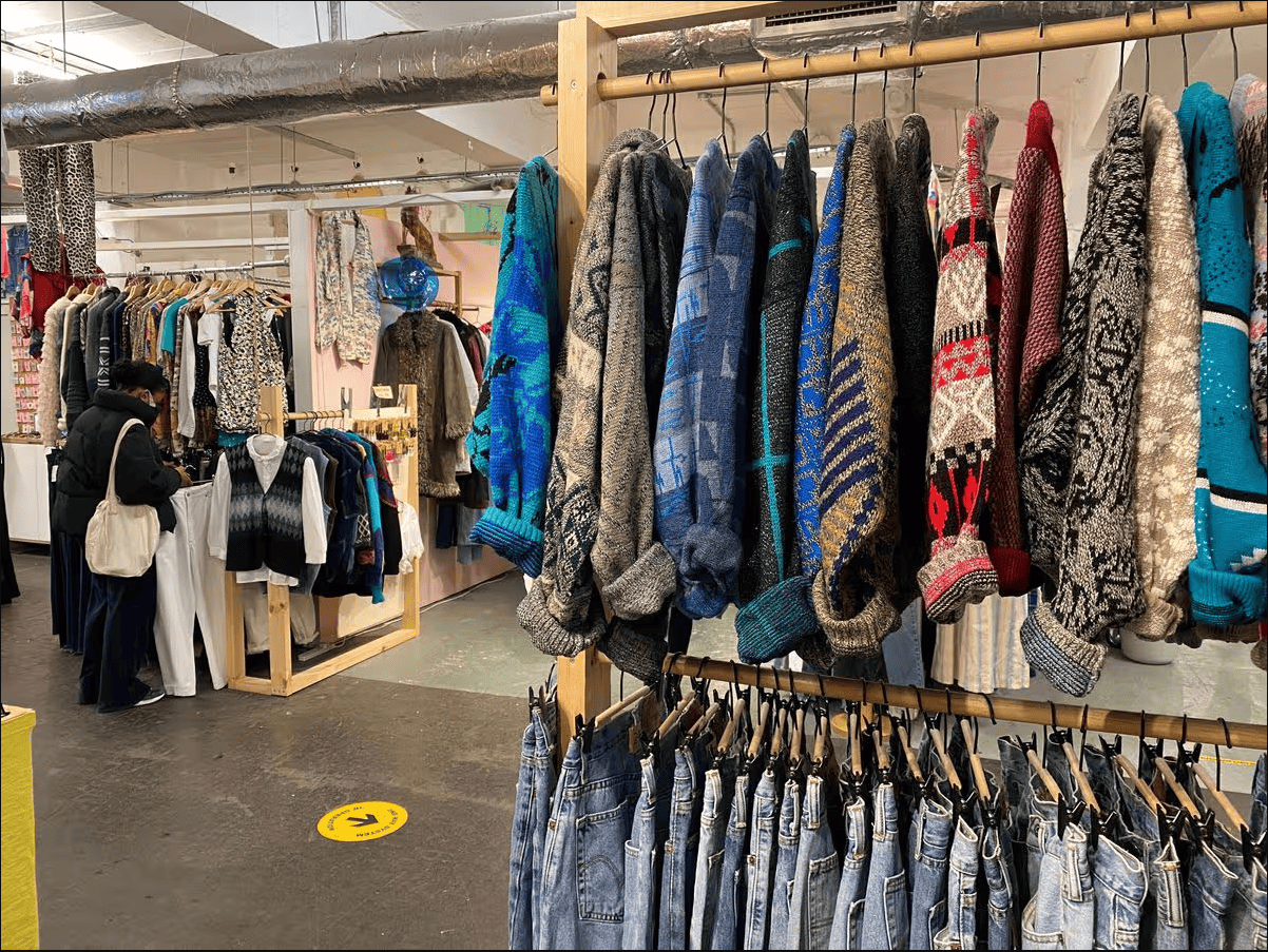 A shopper browses racks of colorful vintage sweaters and denim jeans at an indoor market.