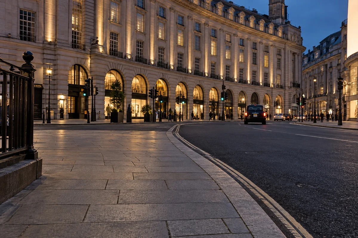 Dry Piccadilly Circus paving and an oblique kerb lead past a dark carriageway toward a lit curved stone fa\u00e7ade.
